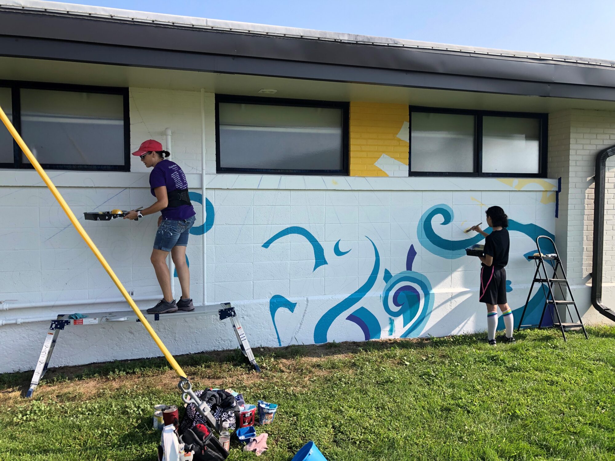 An image of the state of the mural on August 9, 2023 showing the primed wall, the outline of the mural and local artist Theresa Jewell and art student Kaya Munoz beginning to paint in waves.